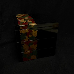 Puede incluir: Una caja bento japonesa negra de varios niveles con un acabado brillante. El lateral de la caja está decorado con un patrón de hojas de otoño en rojo, verde y dorado. La caja está diseñada para contener alimentos.
