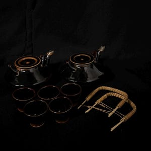 以下が含まれることがあります： 黒い陶器のティーセット。ティーポット2つ、カップ6つ、編み込みの取っ手2つが含まれています。ティーポットには蓋と注ぎ口があります。カップは小さくて丸いです。取っ手は編み込まれた藁でできています。