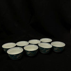 以下が含まれることがあります： 8つの小さくて丸いセラミックボウル。ボウルは白い内側と、花柄のターコイズブルーの外側です。各ボウルの縁を細い青い線が囲んでいます。ボウルは黒い表面に配置されています。