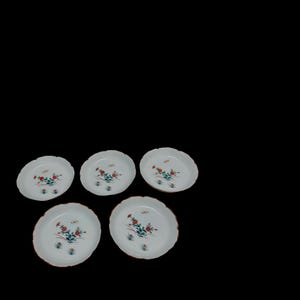 Puede incluir: Cinco platos de cerámica blanca con bordes festoneados. Cada plato presenta un diseño floral con detalles en rojo, azul y verde. Los bordes tienen un borde rojo. Los platos están dispuestos sobre un fondo negro.