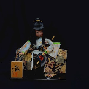 以下が含まれることがあります： 伝統的な衣装を着た日本人形。黒いかつらと、金、黒、白の花柄と幾何学模様の着物。人形は小さな扇子を持ち、日本語の文字が書かれた木箱が添えられています。