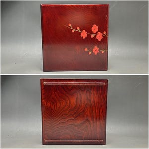 K&ouml;nnte beinhalten: Rechteckige, dunkelrote Holzbox mit floralem Design. Die Oberseite der Box zeigt einen Zweig mit roten Bl&uuml;ten. Die Unterseite der Box hat ein Tablett-&auml;hnliches Design. Die Holzmaserung ist sichtbar.