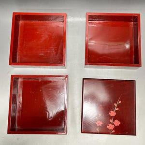 K&ouml;nnte beinhalten: Vierteiliges Set aus rot lackierten Holzkisten. Drei offene Boxen sind quadratisch mit gl&auml;nzendem Finish. Die vierte Box ist ein Deckel, ebenfalls quadratisch, mit dunkelrotem Hintergrund und einem rosa Blumendesign.