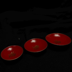 以下が含まれることがあります： 金色の中国の文字が入った、赤色の楕円形のお皿が3つ。お皿は光沢があり、細い金の縁取りがあります。背景は黒です。
