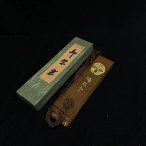 以下が含まれることがあります： 日本の祈りの帯と緑色の箱のセット。祈りの帯は茶色で金色のアクセントと文字が入っています。箱は緑色で、ベージュのラベルに日本語の文字が書かれています。帯は茶色の紐で結ばれています。