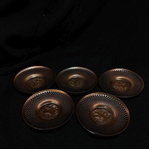 以下が含まれることがあります： ブロンズ色の装飾用ソーサー5枚。穴あきデザインで、それぞれ中央にエンボス加工のデザインが施されています。おそらく花柄または植物柄です。暗い背景に配置されています。
