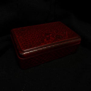 Op de afbeelding: Een rechthoekige, rode sieradendoos met een getextureerd oppervlak en een bloemenontwerp op het deksel. De doos is gemaakt van een donkerrood materiaal en heeft een glanzende afwerking. De doos is gesloten en staat op een donkere achtergrond.