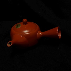 May include: A red ceramic teapot with a round body, a small lid, and a long, flared spout. The teapot has a small handle and a small round knob on the lid. A small sticker is on the top of the teapot. The teapot is on a black background.