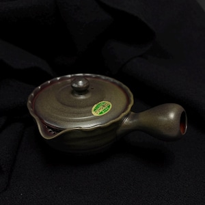 May include: A ceramic teapot with a handle and a lid. The teapot is a muted green color with a dark red interior and a small sticker on the side. The handle is curved and the spout is small.