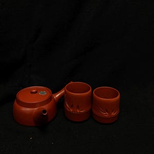 Puede incluir: Un juego de té de cerámica marrón rojizo, que incluye una tetera con asa lateral y dos tazas a juego. Las tazas y la tetera tienen un diseño texturizado. La tetera tiene una tapa pequeña y redonda y un pico. El juego se muestra sobre un fondo oscuro.