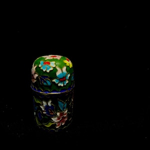 以下が含まれることがあります： 緑色の背景と花の模様が施された装飾的な指ぬき。指ぬきは、金色のアクセントが付いた青、赤、黄色の花のパターンが特徴です。指ぬきの底は青色で、指ぬきは黒い表面に反射しています。