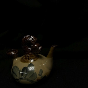 Può includere: Una teiera in ceramica con una testa di animale marrone e strutturata sulla parte superiore. La teiera è beige con disegni floreali blu e verdi. Una piccola tazza è attaccata al lato e un beccuccio si estende dalla parte anteriore.