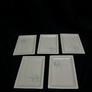 Puede incluir: Cinco platos rectangulares de cerámica blanca con un diseño floral azul claro. Los platos están dispuestos sobre una superficie negra. Cada plato tiene un borde ligeramente elevado y un delicado patrón floral estilizado en la esquina.
