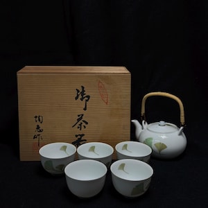 Op de afbeelding: Een witte keramische theeset met een theepot, vier theekopjes en een houten kist. De theepot heeft een bamboe handvat en groene bladeren. De theekopjes hebben ook groene bladeren. De houten kist heeft Japanse karakters.