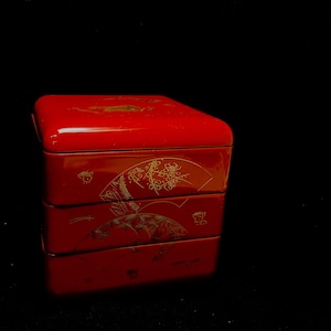 May include: A stackable, red lacquered Japanese bento box. The box has a glossy finish and is decorated with gold-colored fan and floral designs. The box is composed of three stacked compartments with a square lid.