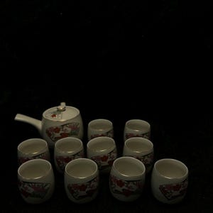 Könnte beinhalten: Ein weißes Keramik-Teeservice mit einer Teekanne und zehn passenden Tassen. Jedes Stück hat einen goldenen Rand und ein dekoratives Blumen- und Fächerdesign in Rot und Rosa. Das Set steht vor einem schwarzen Hintergrund.