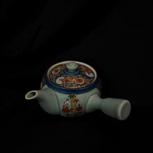 May include: A ceramic teapot in light blue, with a matching lid. The lid is decorated with a floral pattern in red, white, and gold against a blue background. The teapot has a side handle and a spout.