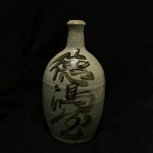 May include: A light-colored ceramic bottle with a narrow neck and rounded body. The bottle features dark brown Japanese calligraphy. The bottle has a slightly textured surface and a matte finish. The background is black.
