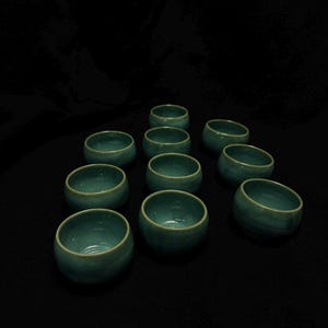 以下が含まれることがあります： 11個の小さなティール色のセラミックボウルのコレクション。各ボウルは丸みを帯びた形状で、わずかに広がった縁があります。ボウルは暗い表面に配置されており、視覚的に魅力的なディスプレイを作成しています。ティール色はボウルにユニークな外観を与えます。
