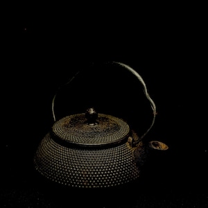 Puede incluir: Tetera de hierro fundido de color marrón oscuro con superficie texturizada y asa curva. La tetera tiene tapa con pomo pequeño y boquilla. Fondo negro.