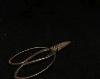 May include: Antique metal scissors with oval finger holes and pointed blades. The scissors have a rusty, aged appearance, suggesting they are vintage or have been well-used. The blades are slightly open, and the scissors are set against a dark background.
