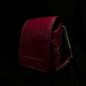 Könnte beinhalten: Ein tief magentafarbener Randoseru-Rucksack mit glänzender, glatter Oberfläche und einer strukturierten, kastenförmigen Silhouette. Er verfügt über eine große Frontklappe mit sichtbaren Nähten, zwei Metallnieten an der Unterseite und verstellbare Schultergurte mit hellem Innenfutter.