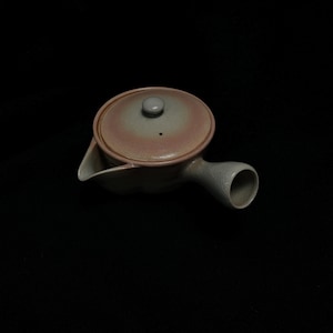 May include: A ceramic side-handled teapot featuring a round body with a soft gradient of muted pink and grey tones. The teapot includes a matching circular lid with a small central knob and a single spout, showcasing a smooth, glazed finish.