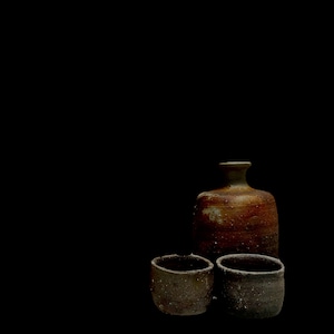 Peut inclure: Une bouteille en céramique rustique avec un vernis texturé, tacheté de brun et d'orange, est placée derrière deux petites tasses cylindriques en céramique gris foncé. Les pièces présentent un aspect terreux et usé avec des détails granulaires.