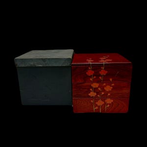 Op de afbeelding: Een zwarte vierkante doos met een getextureerd deksel staat naast een rode houten doos met drie niveaus. De rode doos heeft een oranje en geel bloemenpatroon, met een zichtbaar houtnerfpatroon.