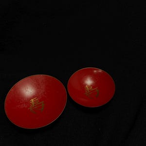 以下が含まれることがあります： それぞれに金色のシンボルが付いた、2つの赤い丸いオブジェクト。オブジェクトは黒い背景に配置されています。オブジェクトは光沢のある素材でできているようです。