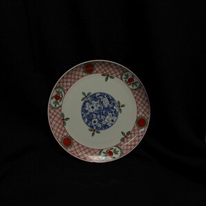 May include: A round, white ceramic plate with a decorative design. The plate features a red and white patterned border, a blue floral centre, and red and green accents. This plate is suitable for serving food.