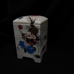 Op de afbeelding: Een witte, driedelige Japanse bento-box van keramiek met een deksel. De doos heeft een handgeschilderde illustratie van een figuur in traditionele kleding, die een rode bal vasthoudt. Het ontwerp bevat een rode zon en blauwe accenten.