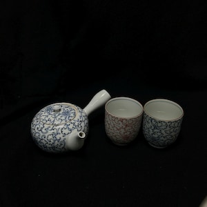 May include: A white ceramic teapot with a side handle and two matching cups. The teapot and one cup feature a blue floral design, while the other cup has a red floral pattern. The set is ideal for serving tea.