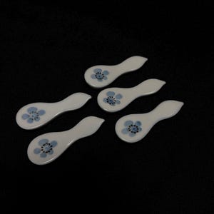 以下が含まれることがあります： 青い花の模様が施された白いセラミック製のスプーンレストが5つ。各レストは、湾曲したハンドルと小さく浅いボウルを備えています。花の模様は、濃い青色の中心を持つ青い花が特徴です。