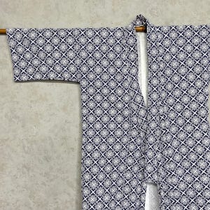 Puede incluir: Un kimono con un patrón geométrico en azul marino y blanco. El kimono está colgado de una barra de madera sobre un fondo gris claro. El patrón consiste en formas de cruces y cuadrados repetidos.