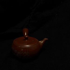以下が含まれることがあります： 蓋と注ぎ口が付いた、小さな茶色の陶器のティーポット。 ティーポットは蓋に取っ手があり、側面に装飾的なデザインが施されています。 ティーポットは暗い背景に置かれています。
