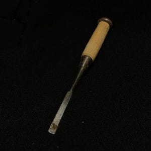 Puede incluir: Un formón para madera con mango de madera clara, una virola de metal oscuro y una hoja de acero plana, larga y estrecha. La herramienta tiene una empuñadura cilíndrica y un eje metálico que se estrecha hacia un filo cortante y rectangular.