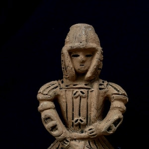 Puede incluir: Pequeña figura de terracota de una persona con armadura. La figura tiene un casco, protectores de hombros y una túnica con elementos decorativos. Los brazos están cruzados y la cara tiene rasgos sencillos. El fondo es negro.
