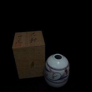 以下が含まれることがあります： 青とピンクの花柄で装飾された、暗い開口部のある白いセラミック花瓶。 花瓶は卵形で、日本語の文字が入った木箱の隣にあります。 背景は黒です。