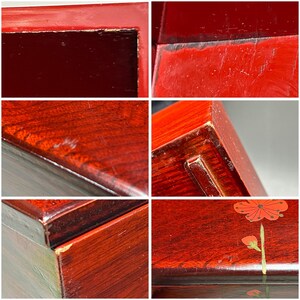 K&ouml;nnte beinhalten: Nahaufnahmen einer lackierten Holzkiste im Vintage-Stil. Die Kiste ist tiefrot und weist sichtbare Gebrauchsspuren auf, darunter Kratzer und abgebrochene Kanten. Eine Platte zeigt eine bemalte rote Blume mit gr&uuml;nen Akzenten.