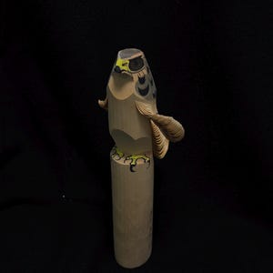 Könnte beinhalten: Eine geschnitzte Holzskulptur eines Falken. Der Falke hat einen hellbraunen Körper mit schwarzen und grauen Markierungen auf Kopf und Flügeln. Die Krallen des Falken sind grün und gelb bemalt. Die Skulptur ist auf einem zylindrischen Holzsockel montiert.