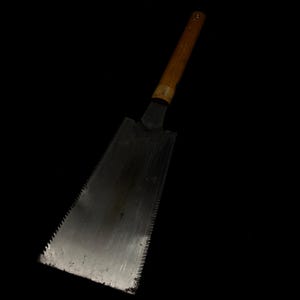 May include: A Japanese pull saw with a wooden handle and a metal blade. The blade is rectangular with a toothed edge for cutting. The handle is a light brown color.