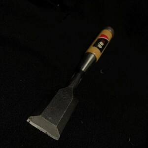 May include: A Japanese wood chisel featuring a wide, flat, dark metal blade with a beveled edge and a light-colored wooden handle. The handle includes a metal ferrule at the top and a black label with white Japanese characters and a red accent.