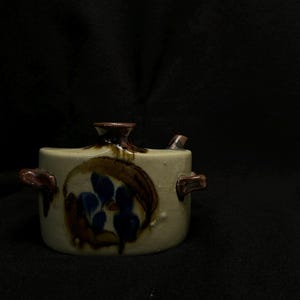 May include: A ceramic creamer with a cream-colored base, adorned with brown and blue floral designs. It features a spout, a handle on each side, and a dark brown top. The creamer is set against a black backdrop.