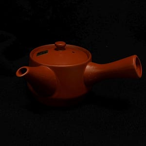 May include: A reddish-brown ceramic teapot with a lid and a handle. The teapot has a spout and a small sticker on the side. The design is simple and functional, suitable for brewing tea. The teapot is set against a dark background.