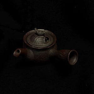 May include: A dark brown teapot with a bird-shaped handle and a bird figurine on the lid. The teapot has a spout and a handle, and it appears to be made of ceramic or a similar material. The design is detailed and textured.