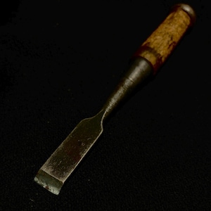 May include: A vintage wood chisel with a metal blade and a textured wooden handle. The handle is a light brown colour with darker brown accents. The blade is a dull silver colour.