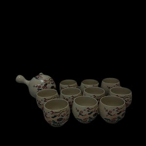 Könnte beinhalten: Cremefarbenes Keramik-Teeservice mit einer Teekanne und zehn passenden Tassen. Die Teekanne hat einen gebogenen Griff und einen Deckel. Die Tassen und die Teekanne sind mit einem Blumen- und Wolkenmuster in Rot, Blau und Gold verziert.