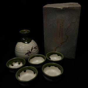 May include: A Japanese sake set. The white ceramic bottle has a green top and bamboo design, with five small matching cups with green rims. A textured box with Japanese text is in the background.