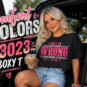 Może przedstawiać: Czarny krótki t-shirt z napisem "I MAY BE WRONG but I doubt it" w kolorze różowym i białym. Obraz zawiera również tekst "Comfort COLORS 3023 BOXY T". Osoba trzyma filiżankę kawy.
